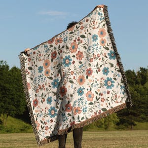 May include: A white woven throw blanket with a floral pattern in shades of blue, orange, and brown. The blanket has a fringed edge.