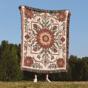 Puede incluir: Una manta tejida de color crema con un dise&ntilde;o floral geom&eacute;trico en tonos marrones, verdes y amarillos. La manta tiene un borde con flecos y se sostiene contra un fondo de cielo azul.