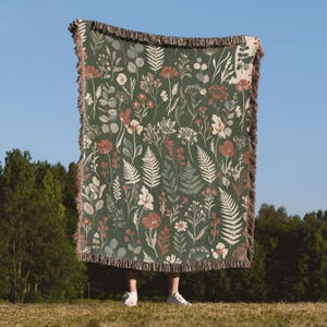 May include: A rectangular woven throw blanket featuring a dark green background with a repeating pattern of various botanical elements including ferns, wildflowers, and eucalyptus leaves in shades of cream, rust orange, and muted blue, with fringed edges.