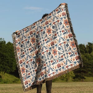 May include: A patterned throw blanket featuring a floral and heart design in brown, blue, and peach hues on a white backdrop. The blanket, with a fringed border, is held up against a blue sky.