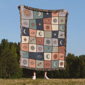 May include: A woven throw blanket featuring a grid pattern of celestial motifs including suns, moons, planets, stars, and clouds. The design uses a muted colour palette of blue, cream, and terracotta, finished with decorative fringe on all edges.