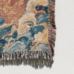 May include: A woven throw blanket with a detailed illustration of swirling waves in shades of blue, tan, and peach. The blanket has a fringed edge with a mix of colors. The design evokes a sense of movement and artistic flair.
