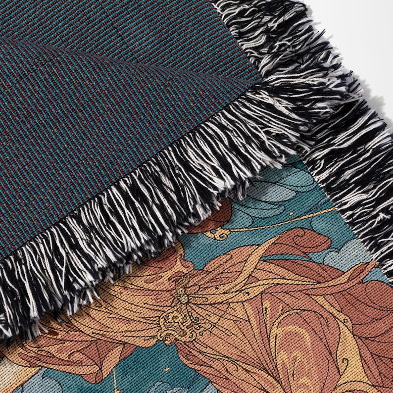 May include: Close-up of a woven throw blanket with a detailed design. The blanket features a teal and maroon textured top layer, and a bottom layer with a floral and cloud pattern in shades of orange, yellow, and blue. The edges are finished with black and white fringe.