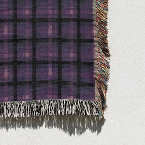 May include: A close-up of a purple plaid throw blanket with black grid lines. The blanket has a fringed edge with a mix of colorful threads. The blanket is on a white background.