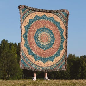 May include: A large, round woven blanket with a mandala design in shades of beige, teal, and coral. The blanket has a fringed edge and is held up against a blue sky background. The design features intricate floral and geometric patterns.