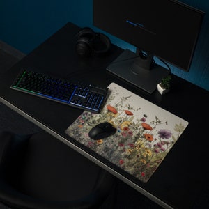 May include: A black keyboard with rainbow lighting sits on a black desk with a floral mousepad. A black computer mouse is on the mousepad. A pair of black headphones and a small potted plant are also on the desk.