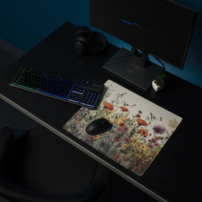 May include: A black keyboard with rainbow lighting sits on a black desk with a floral mousepad. A black computer mouse is on the mousepad. A pair of black headphones and a small potted plant are also on the desk.