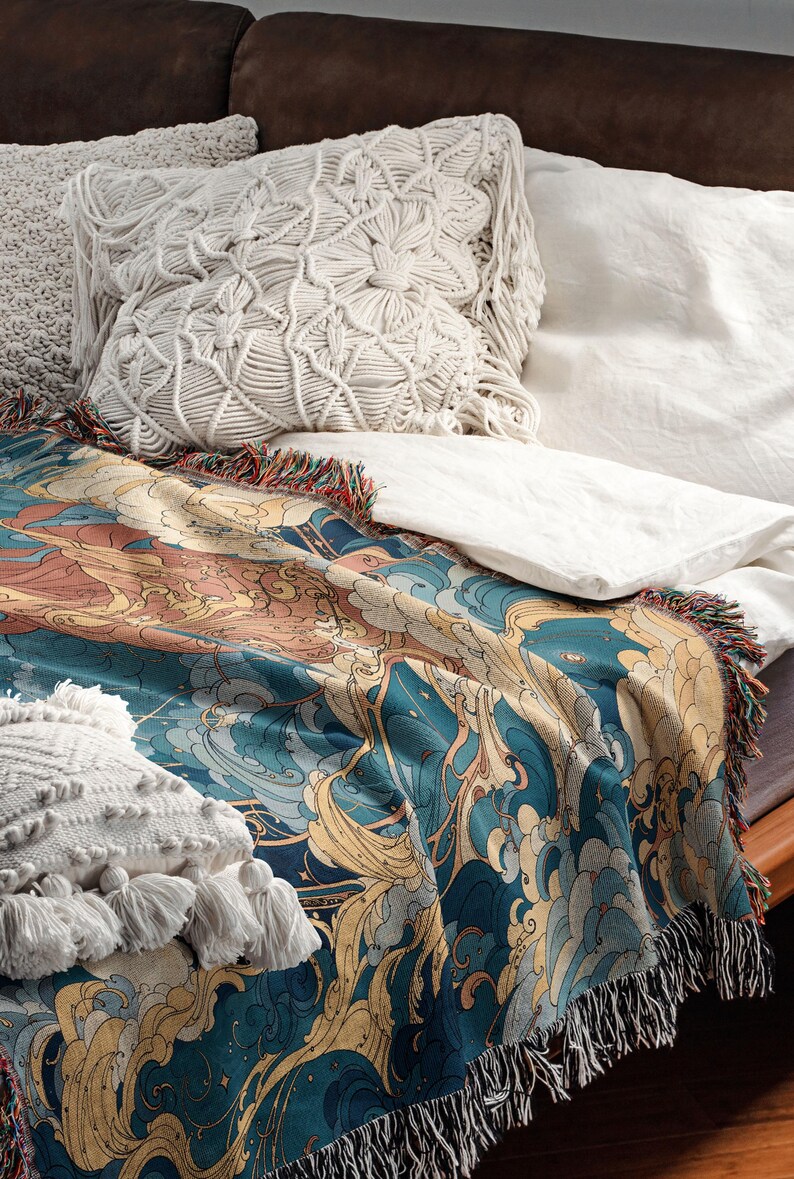 May include: A decorative throw blanket with a blue, gold, and peach wave pattern, fringed edges, and a soft texture. The blanket is draped over a bed with white linens and decorative pillows.