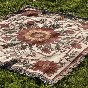 Puede incluir: Una manta cuadrada de color crema con un estampado floral en tonos rojos, marrones y amarillos. La manta tiene un dise&ntilde;o geom&eacute;trico con un motivo floral central y est&aacute; rematada con flecos en los bordes. La manta est&aacute; sobre hierba verde.