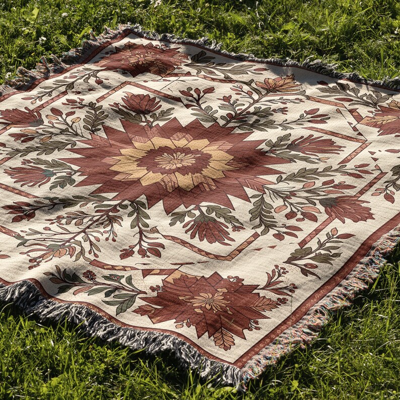 Puede incluir: Una manta cuadrada de color crema con un estampado floral en tonos rojos, marrones y amarillos. La manta tiene un dise&ntilde;o geom&eacute;trico con un motivo floral central y est&aacute; rematada con flecos en los bordes. La manta est&aacute; sobre hierba verde.