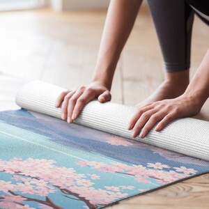 May include: A white yoga mat with a floral design featuring pink cherry blossoms on a blue background. The mat is rolled up and being held by a person's hands.