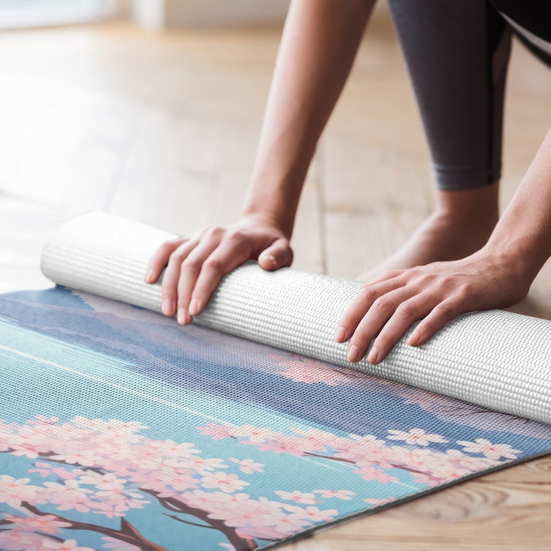 May include: A white yoga mat with a floral design featuring pink cherry blossoms on a blue background. The mat is rolled up and being held by a person's hands.