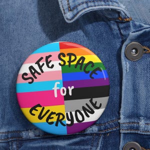 Puede incluir: Un botón redondo con el texto "SAFE SPACE for EVERYONE" en letras negras. El botón presenta un diseño colorido que incorpora varias banderas del orgullo, incluyendo las banderas transgénero, arcoíris y asexual. El botón está sobre una chaqueta vaquera.