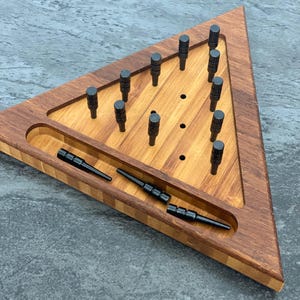 May include: A wooden triangular peg game with a dark brown frame and a lighter wood interior. The game includes black pegs and a recessed area for storing the pegs. The game is set against a gray background.