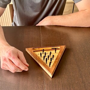 May include: A wooden triangular puzzle game with a light wood interior and a darker wood frame. The game features black pegs and a small channel at the top. The game is set on a dark brown table.