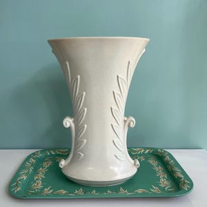 May include: A cream-colored ceramic vase with leaf designs and handles, set on a rectangular green tray with a floral pattern. The vase has a textured surface and a flared top.