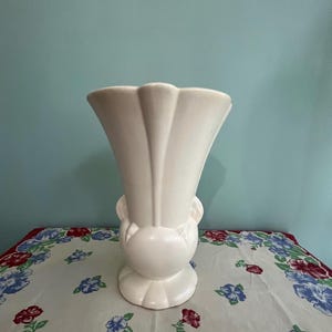 May include: A white ceramic vase with a fluted, fan-shaped top and a rounded base. The vase has a smooth, glossy finish and stands on a floral patterned tablecloth. The vase is approximately 10 inches tall.