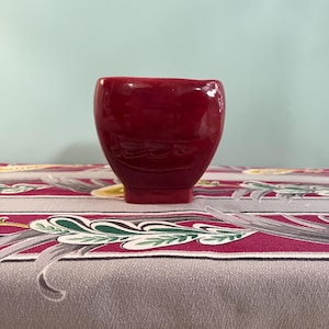 May include: A small, glossy, deep red ceramic bowl with a rounded body and a square base. The bowl is set against a patterned tablecloth with a gray background and floral designs. The background is a light blue.