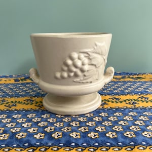 May include: A cream-colored ceramic planter with a pedestal base and two small handles. The planter features a raised relief design of grapes and leaves. The planter is sitting on a blue and yellow patterned tablecloth.