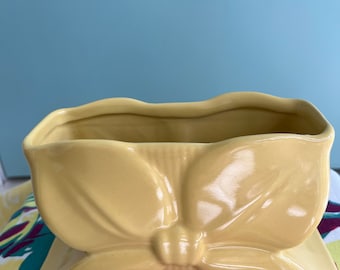 May include: A yellow ceramic planter shaped like a bow. The planter has a smooth, glossy finish and is sitting on a floral fabric.