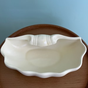 May include: A white ceramic dish with a scalloped edge and a raised handle in the center. The dish is sitting on a wooden surface.