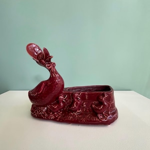 May include: A burgundy ceramic planter featuring a duck design. The planter has a rectangular base with duckling relief details and a tall, curved neck duck figure. The planter is suitable for small plants or flowers.