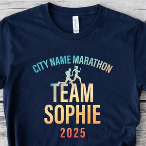 以下が含まれることがあります： 紺色のTシャツ。上部に「CITY NAME MARATHON」の文字、その下にランナーのグラフィック。グラフィックの下には「TEAM SOPHIE 2025」がグラデーションカラーで表示されています。Tシャツは裾で結ばれています。