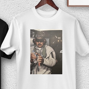 May include: Black and white t-shirts hanging on a wooden hanger. Each shirt features a printed image of a person holding two bottles. The person is wearing a hat, sunglasses, and a gray jacket.