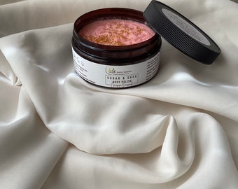 May include: A jar of Sugar & Shea Body Polish with a pink, textured interior and gold flecks. The jar is brown with a black lid resting beside it. The label reads "Champagne Dreams". The jar sits on a white, silky surface.