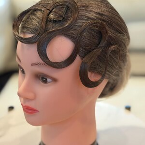 May include: A mannequin head with brown hair styled in prominent, sculpted finger waves across the forehead. The hair is dark brown with lighter highlights, and the mannequin features painted facial details including brown eyes and pink lips.