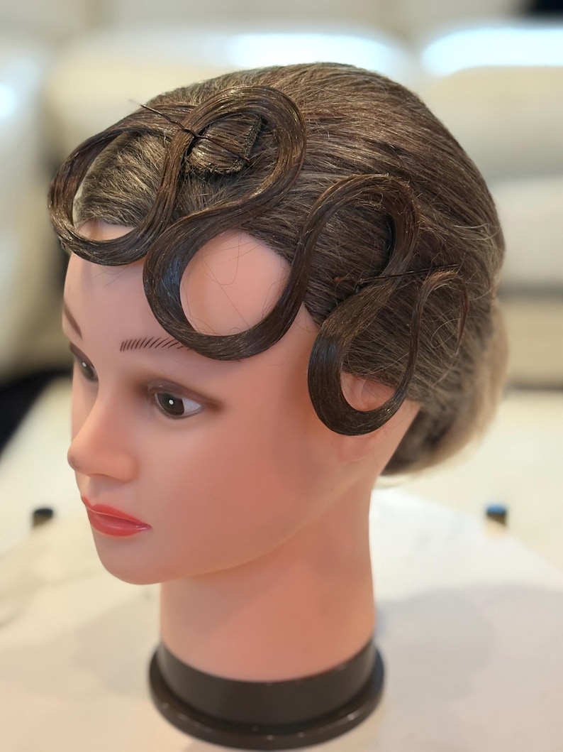 May include: A mannequin head with brown hair styled in prominent, sculpted finger waves across the forehead. The hair is dark brown with lighter highlights, and the mannequin features painted facial details including brown eyes and pink lips.