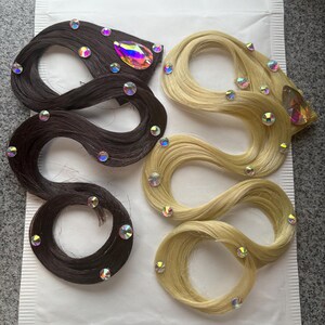 May include: Two hair extensions, one dark brown and one blonde, arranged in wavy shapes. Each extension is decorated with multiple small, round, iridescent rhinestones and one large, teardrop-shaped, multicoloured crystal accent.
