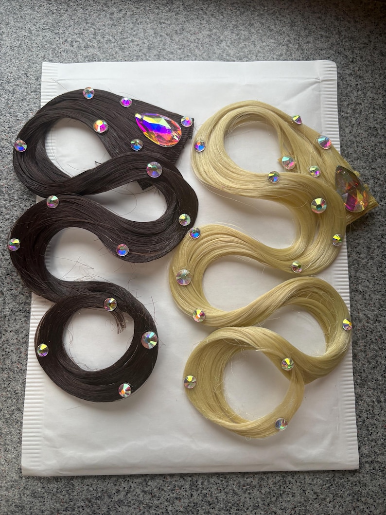 May include: Two hair extensions, one dark brown and one blonde, arranged in wavy shapes. Each extension is decorated with multiple small, round, iridescent rhinestones and one large, teardrop-shaped, multicoloured crystal accent.