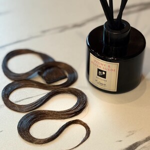 May include: A black cylindrical reed diffuser with a label reading Peony & Blush Suede Diffuser by Deluxe, featuring black reeds, placed next to a long, wavy, dark brown hair extension piece with a small rectangular attachment tab at the top.