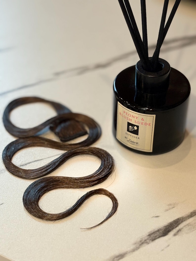 May include: A black cylindrical reed diffuser with a label reading Peony & Blush Suede Diffuser by Deluxe, featuring black reeds, placed next to a long, wavy, dark brown hair extension piece with a small rectangular attachment tab at the top.