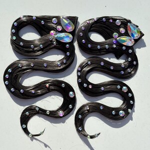 May include: Two hair extensions featuring long, wavy black strands arranged in a serpentine shape. Each piece is embellished with numerous small, round, iridescent rhinestones and two larger, teardrop-shaped iridescent crystals at the top.