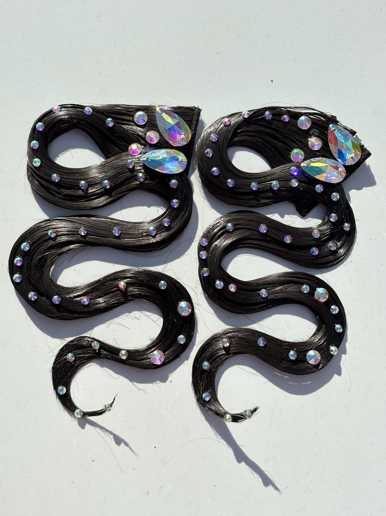 May include: Two hair extensions featuring long, wavy black strands arranged in a serpentine shape. Each piece is embellished with numerous small, round, iridescent rhinestones and two larger, teardrop-shaped iridescent crystals at the top.
