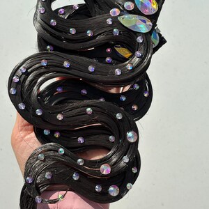 May include: A black hair extension piece styled in wavy strands, embellished with numerous small, round, iridescent rhinestones and two larger, teardrop-shaped, multi-coloured crystals attached near the top edge of the hair section.