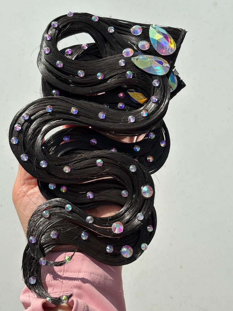 May include: A black hair extension piece styled in wavy strands, embellished with numerous small, round, iridescent rhinestones and two larger, teardrop-shaped, multi-coloured crystals attached near the top edge of the hair section.