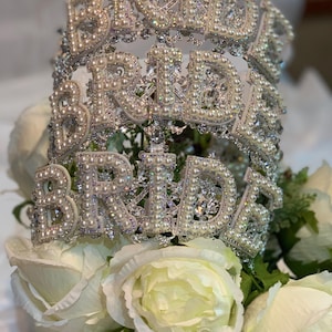 May include: A silver tiara with the word "BRIDE" spelled out in large, pearl-covered letters. The tiara sits atop white roses and greenery, creating a bridal theme.