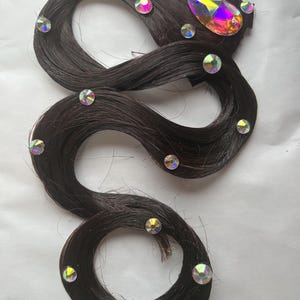 May include: A dark brown hair extension piece arranged in a serpentine shape, adorned with multiple small, round, iridescent rhinestones and one large, teardrop-shaped, multi-coloured iridescent crystal accent attached to the top section.