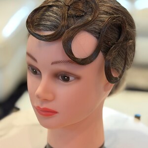 May include: A mannequin head with a smooth, light-toned complexion and painted facial features, including dark eyebrows, brown eyes, and red lips. The head features dark brown hair styled into distinct, flat, wave-like curls across the forehead.