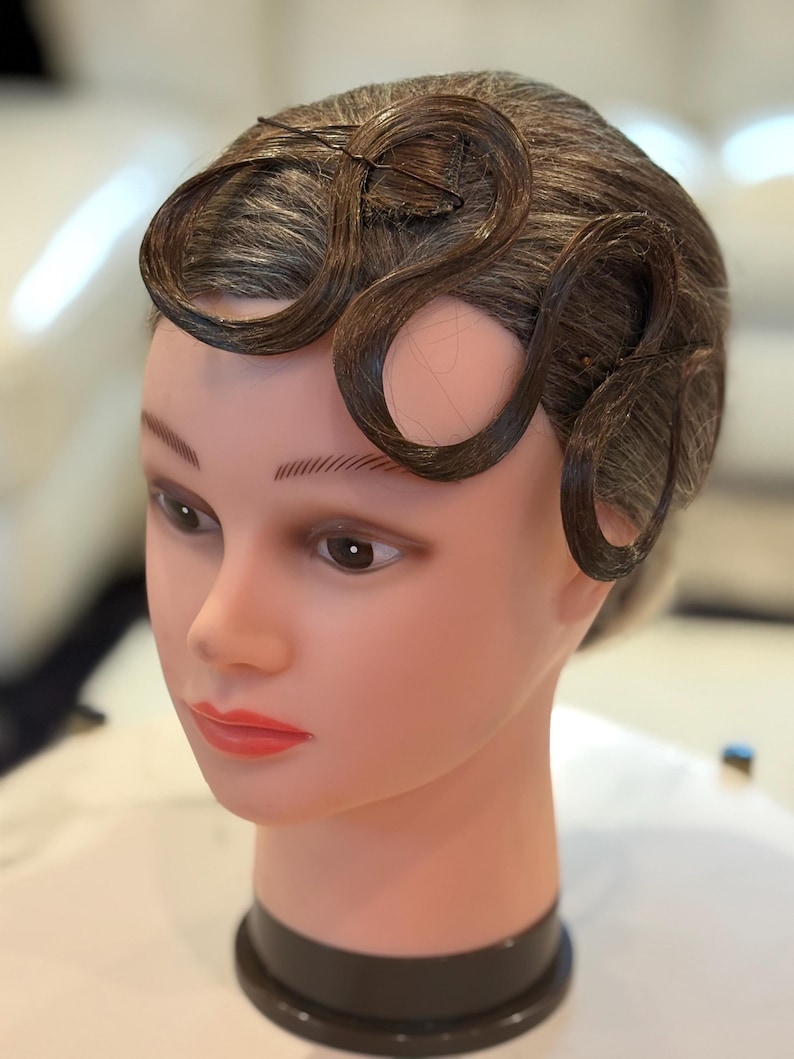 May include: A mannequin head with a smooth, light-toned complexion and painted facial features, including dark eyebrows, brown eyes, and red lips. The head features dark brown hair styled into distinct, flat, wave-like curls across the forehead.