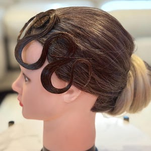May include: A mannequin head featuring a hairstyle with dark brown hair styled into flat, S-shaped finger waves along the temple and forehead, secured with thin hairpins, and a low bun at the back with lighter blonde hair tones visible.