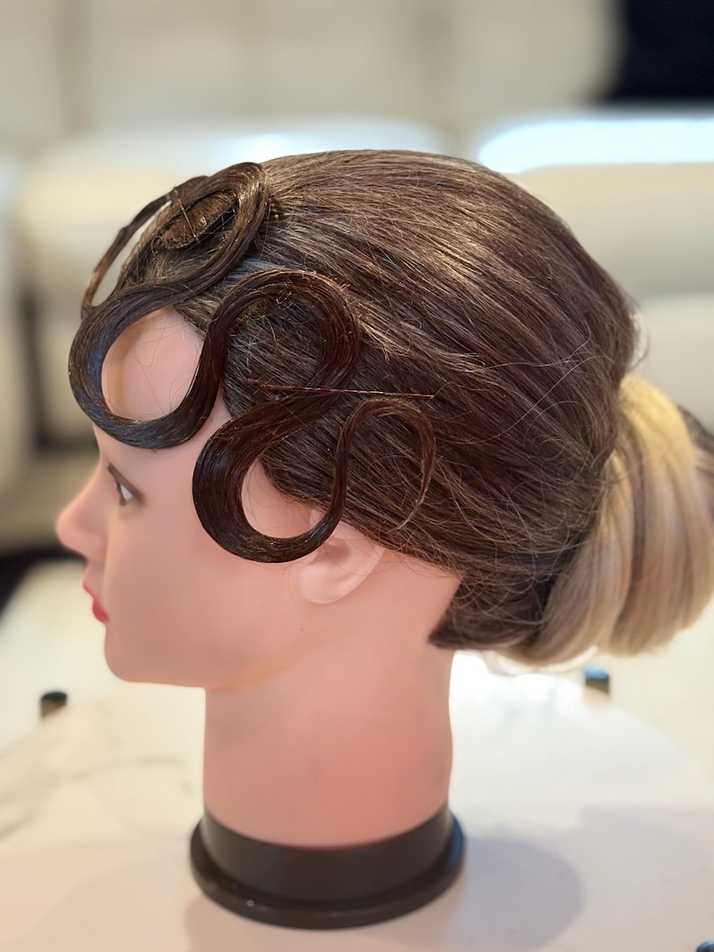 May include: A mannequin head featuring a hairstyle with dark brown hair styled into flat, S-shaped finger waves along the temple and forehead, secured with thin hairpins, and a low bun at the back with lighter blonde hair tones visible.