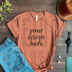 May include: A rust-coloured t-shirt with the text "your design here" printed on the front. The t-shirt is laid flat on a wooden surface with a pair of blue jeans, a brown hat, a black watch, and a pair of white earbuds.