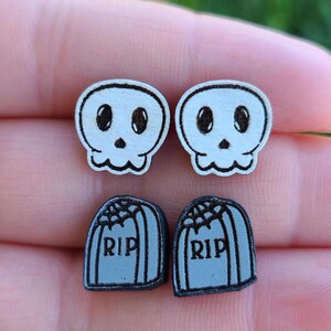 May include: A pair of Halloween-themed earrings featuring two white skull designs and two gray tombstone designs with the word "RIP" on each.