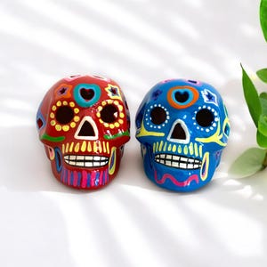 May include: Two colorful sugar skull figurines, one red and one blue, with intricate painted details. The skulls are decorated with yellow, pink, and white accents.