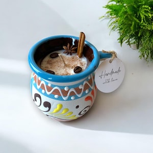 Op de afbeelding: Een blauwe en witte keramische mok-kaars met een kaneelstokje, steranijs en koffiebonen erop. De kaars is versierd met een kleurrijk bloemenpatroon en de woorden "Handmade with love" staan op een wit label dat aan de handgreep is bevestigd.