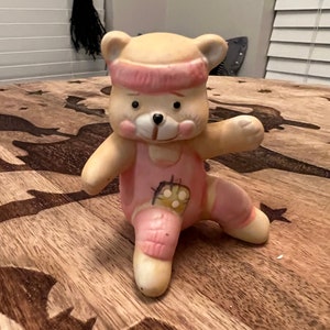 May include: A ceramic figurine of a bear wearing a pink leotard and headband. The bear is standing on a wooden surface with a bat design.